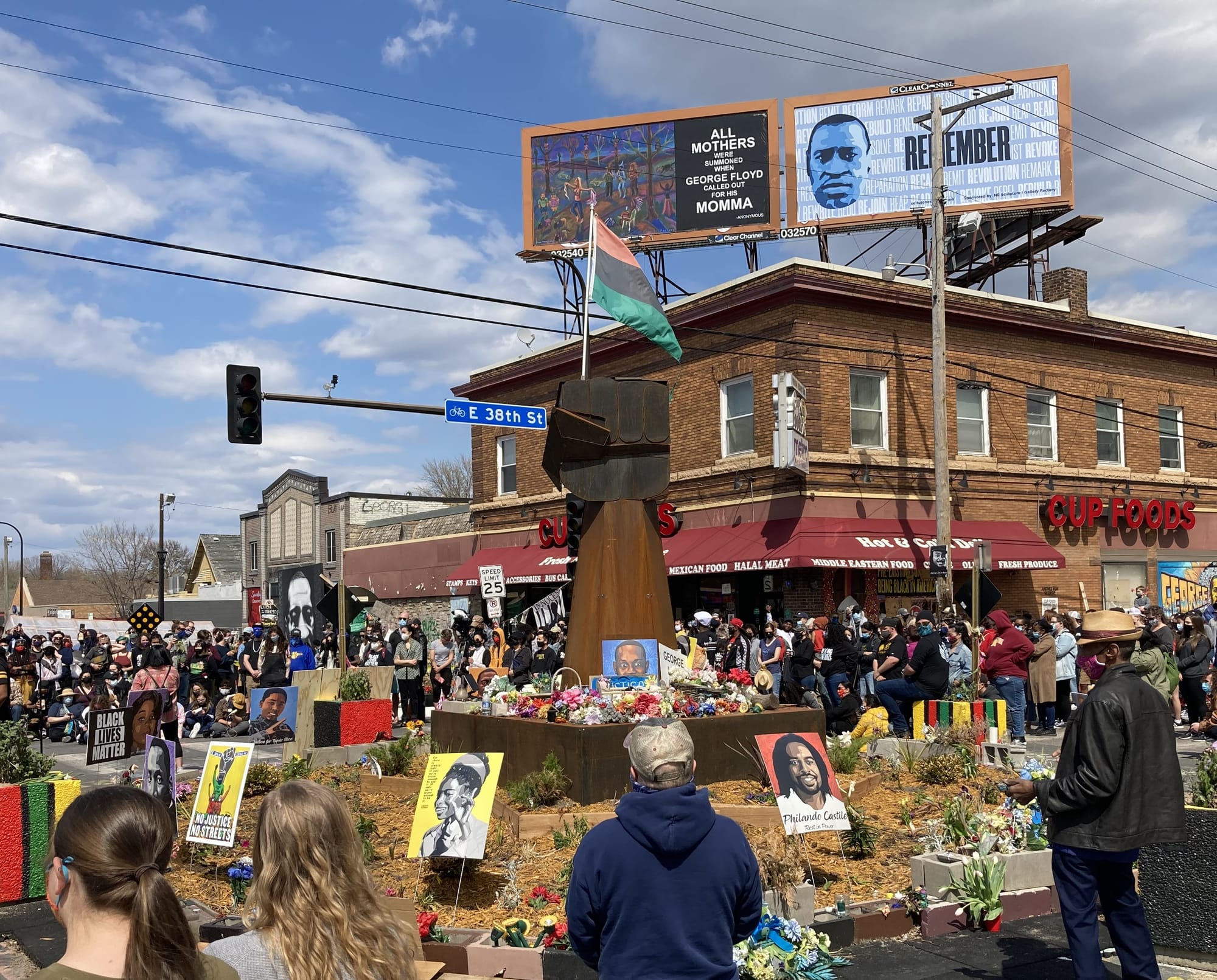 A rust colored fist with a red, black, and green horizontal striped Black Power flag, surrounded by images of victims of police violence, sits in the middle of a street intersection amid hundreds of people.