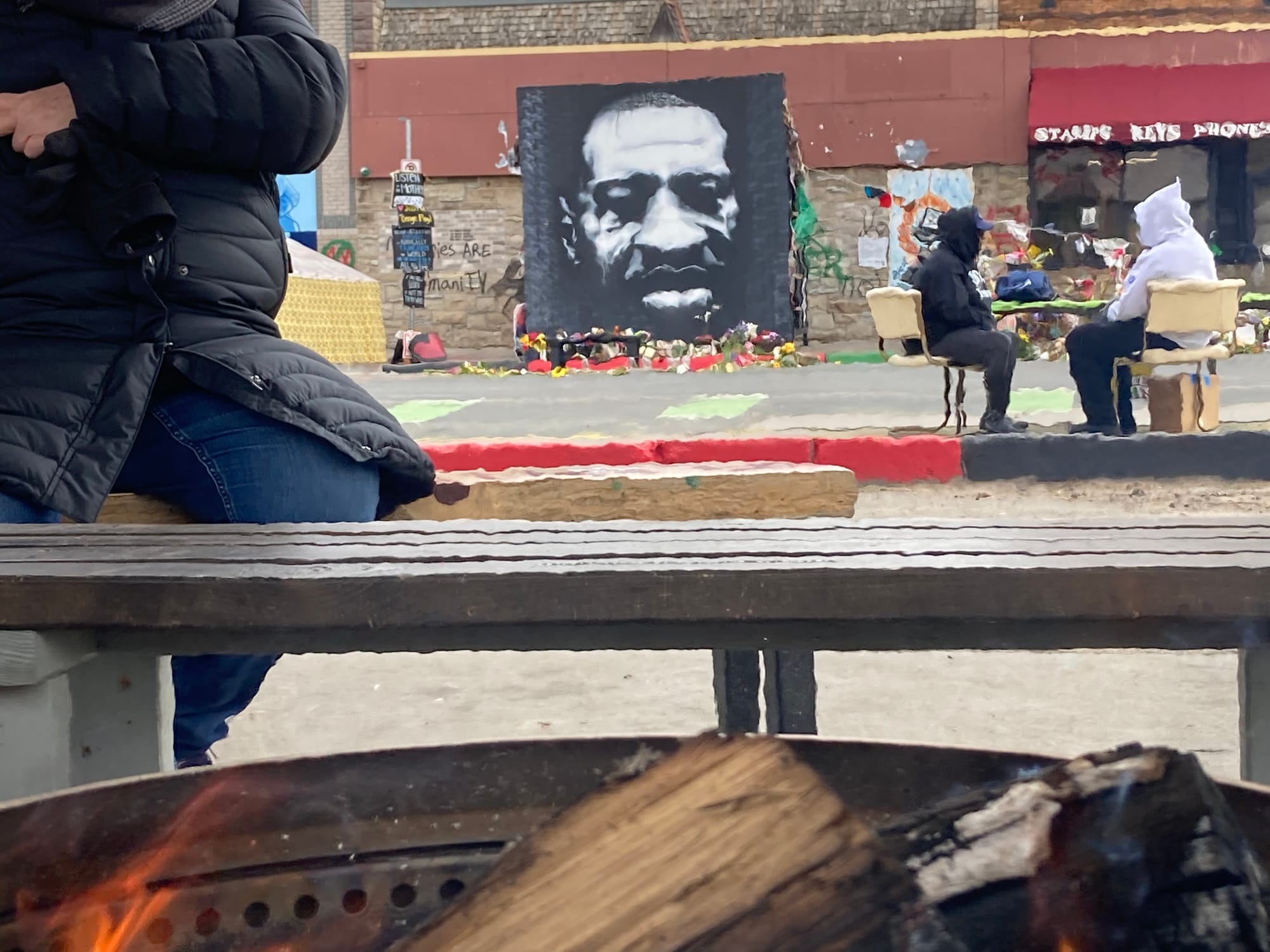 In the foreground, a fire is visible and a person in a heavy coat sits on a bench. In distance, two people sit at table near a black and white painting of George Floyd surrounded by flowers.