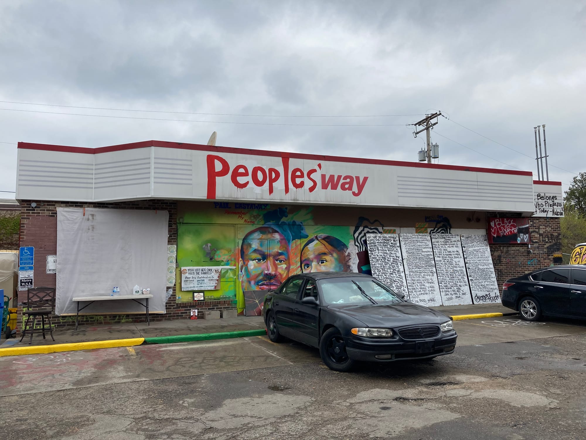 A photo of an abandoned gas station convenience store that has been re-named "People's Way". The building is covered in a mural. A list of demands is written on plywood in front of the building. Cars are parked on the pavement.