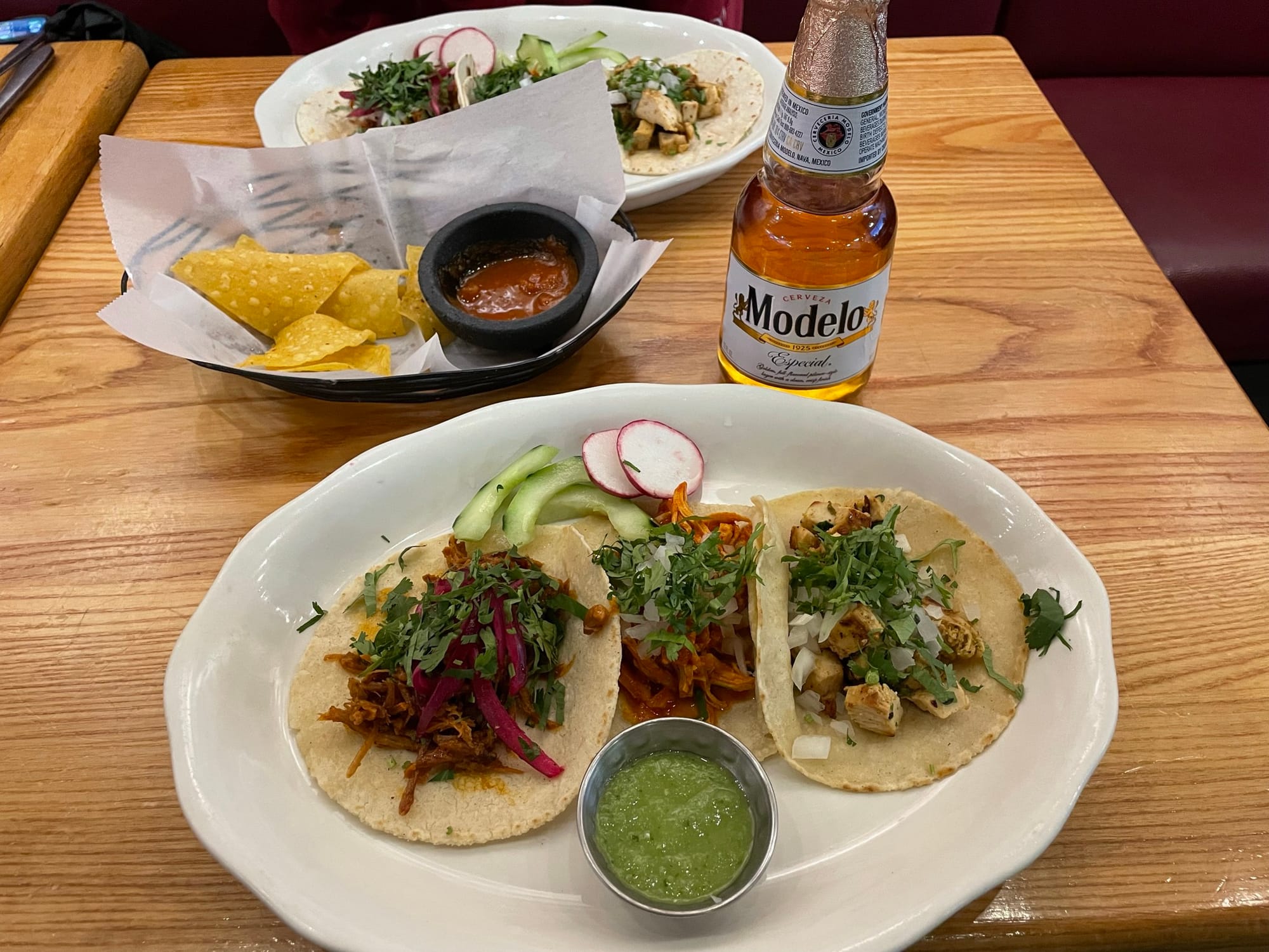 Two plates with three tacos and a salsa, two bottles of Modelo beer, and a basket of chips and salsa sit on a wood table.
